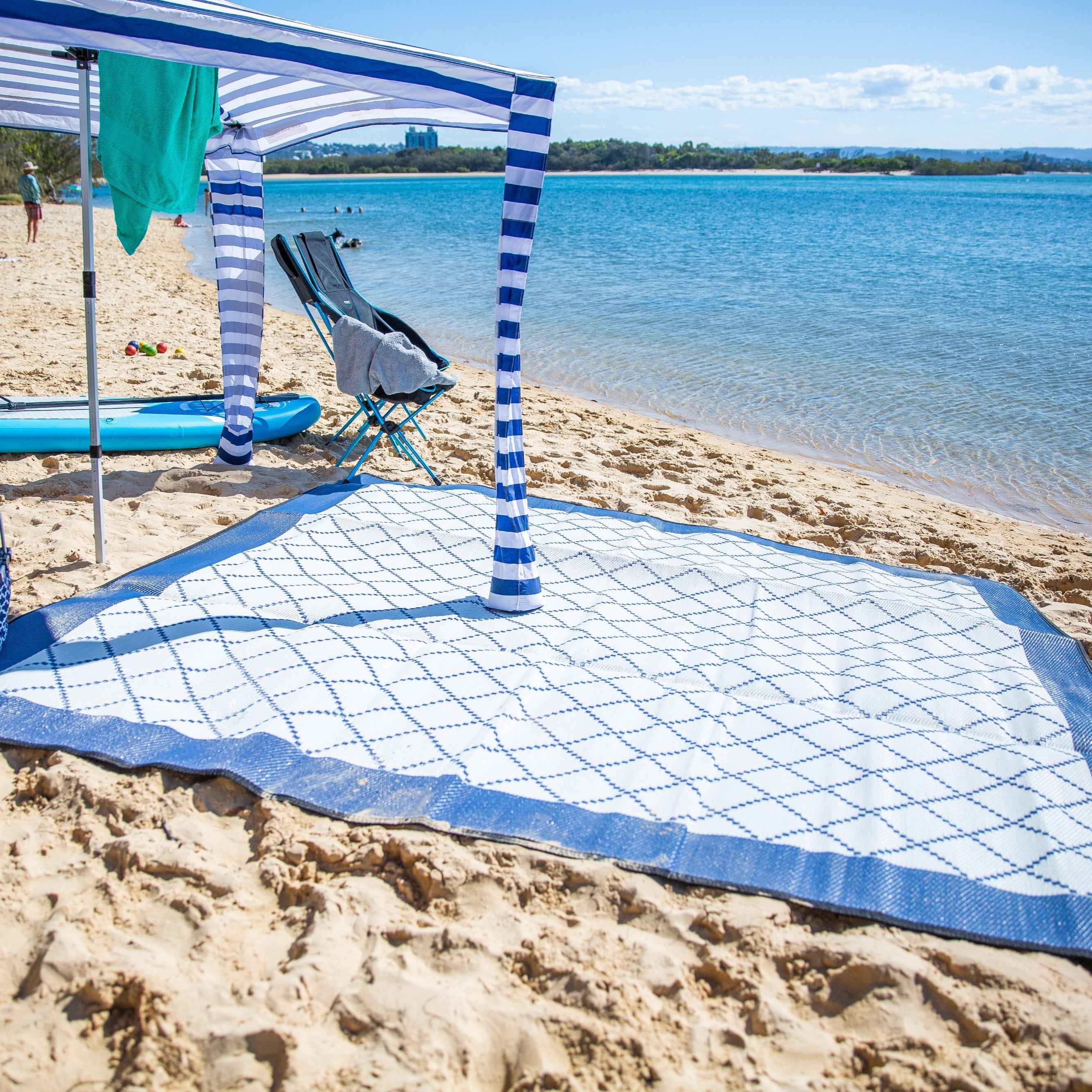 A colorful recycled mat laid out on a sandy beach with gentle ocean waves in the background.
