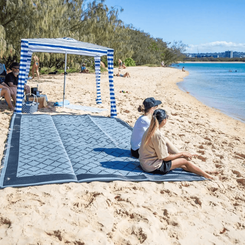 Two people relaxing on a blue patterned outdoor mat at the beach under a striped canopy, perfect for picnics and sand days.