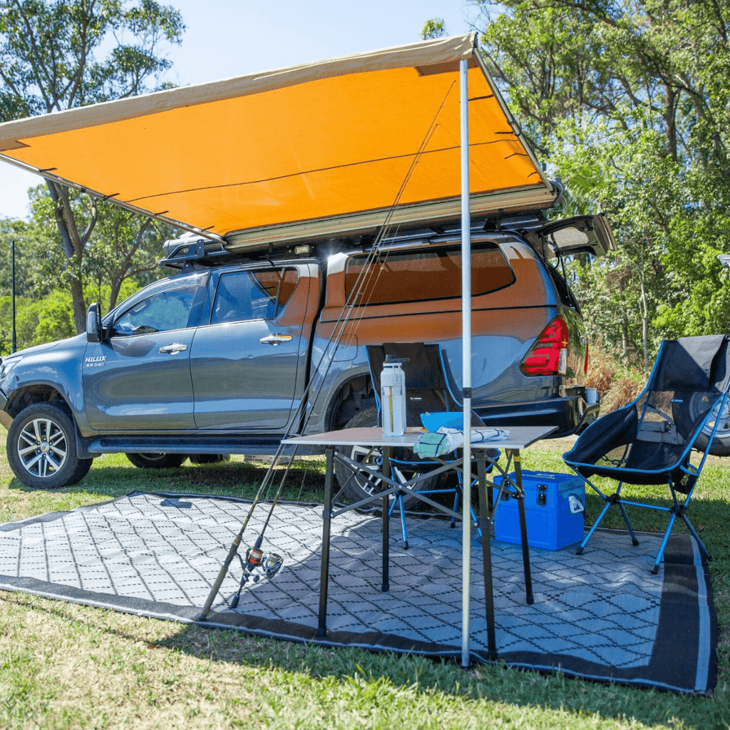 Recycled outdoor mat under camp setup with truck, table, chairs, and awning in grassy area