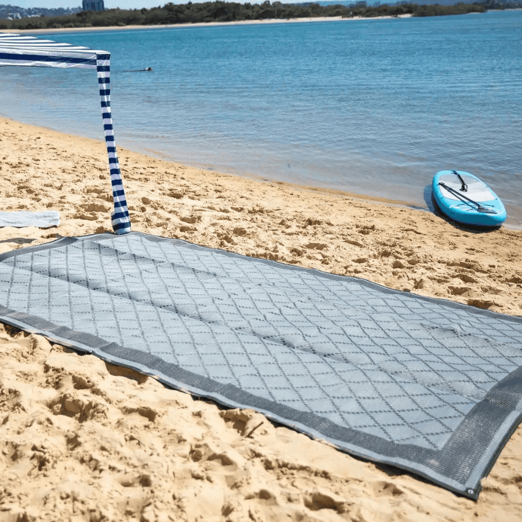 Gray diamond-patterned outdoor mat on sandy beach near blue water, perfect for picnics or beach days