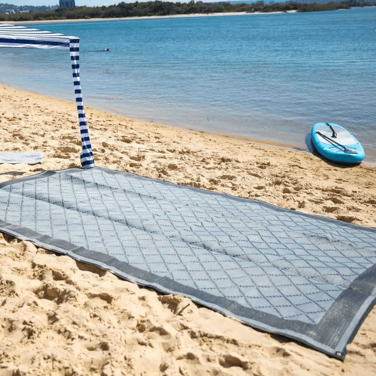 Gray diamond-patterned outdoor mat on sandy beach near blue water, perfect for picnics or beach days