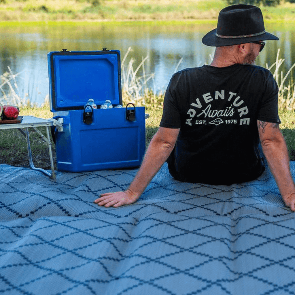 Man relaxing on blue patterned outdoor mat by lake with cooler, perfect for picnics and camping adventures