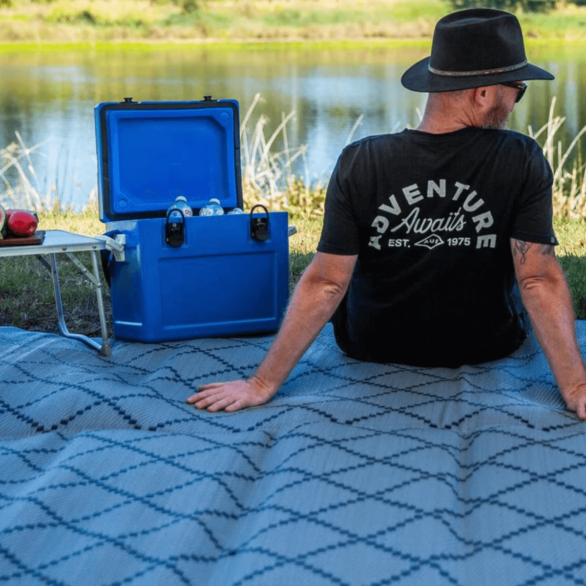 Man relaxing on blue patterned outdoor mat by lake with cooler, perfect for picnics and camping adventures