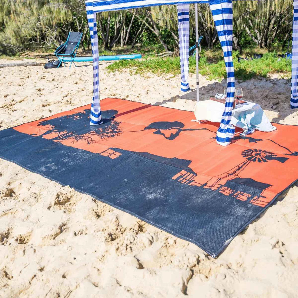 Orange and navy recycled outdoor mat with farm design on sandy beach under striped canopy