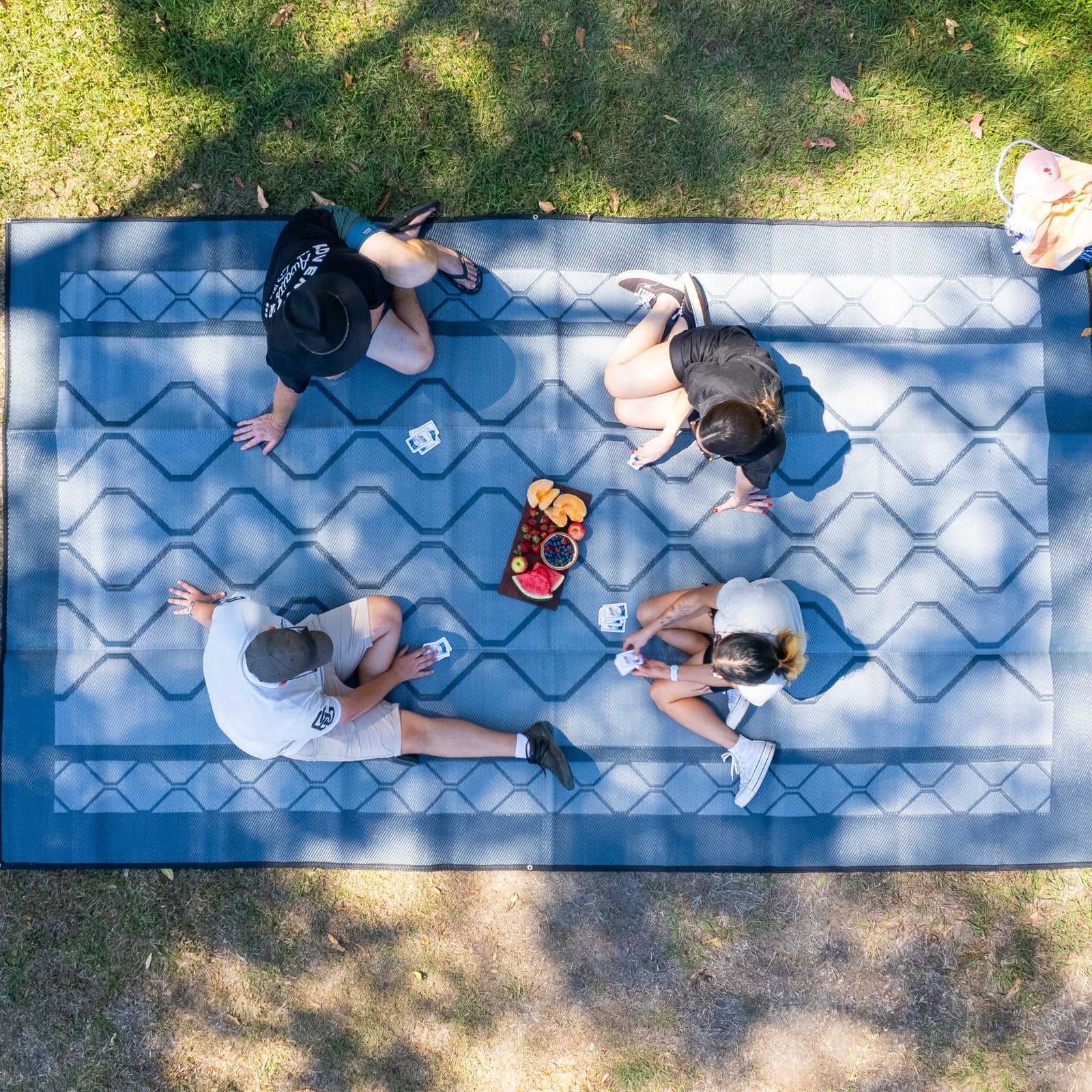 Four people enjoying a picnic on a blue geometric-patterned outdoor mat in grass, perfect for parks and picnics.