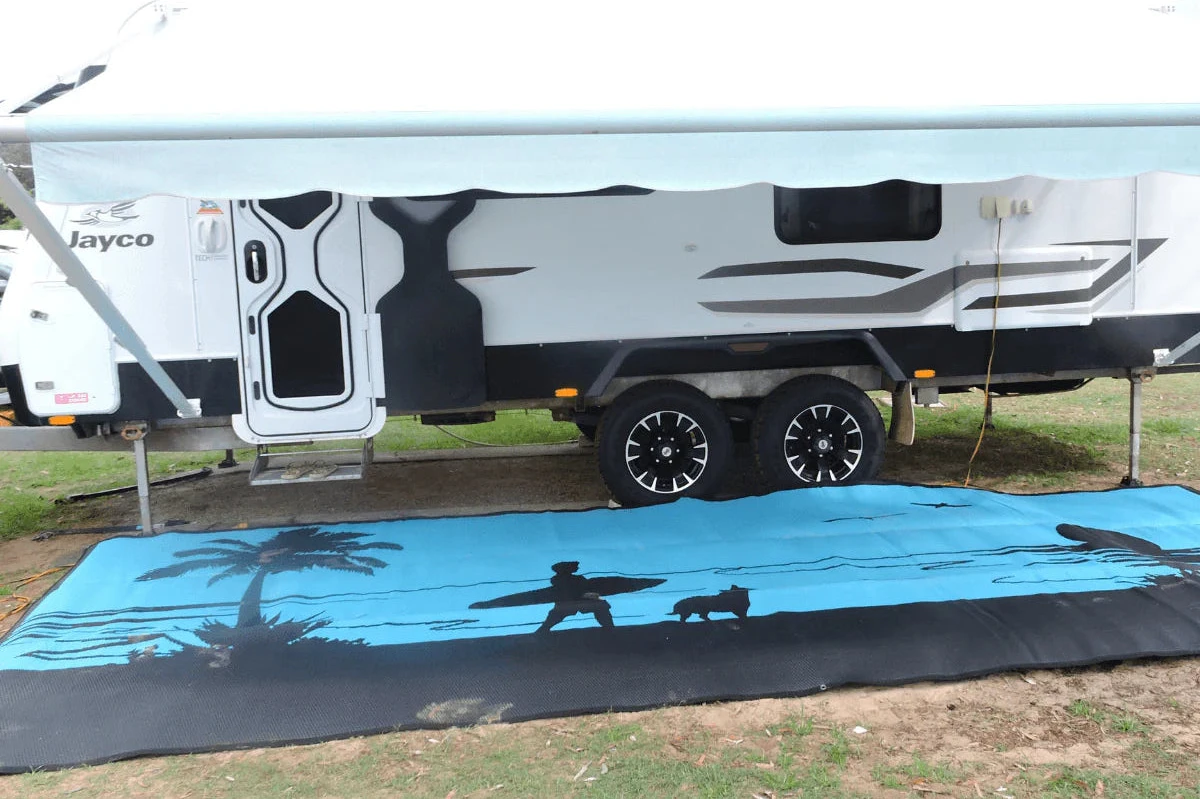 Blue beach mat with surfer and palm tree design under caravan, ideal for camping and outdoor adventures.