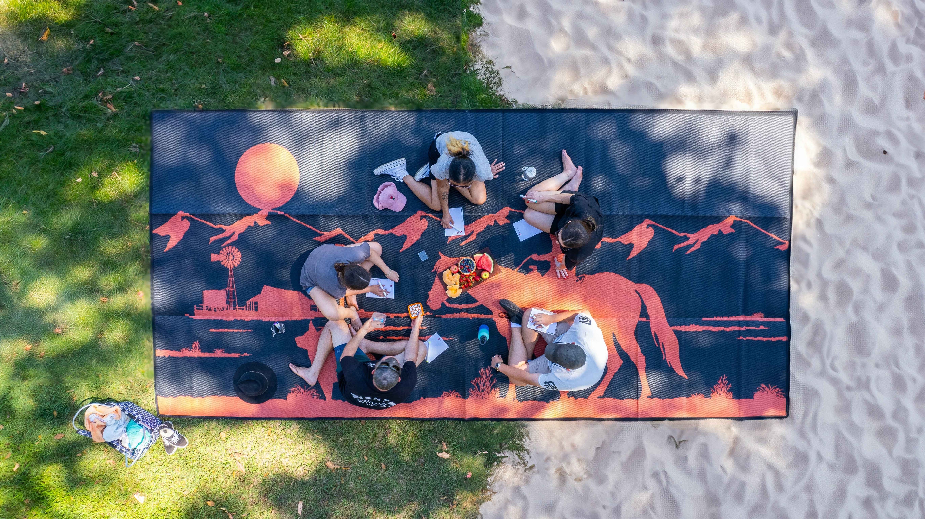 Group enjoying picnic on large outdoor mat with sunset horse design, perfect for beach or park outings.