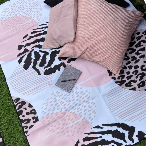Stylish pink and black patterned outdoor mat with pillows and notebook on grass, perfect for picnics or beach days.