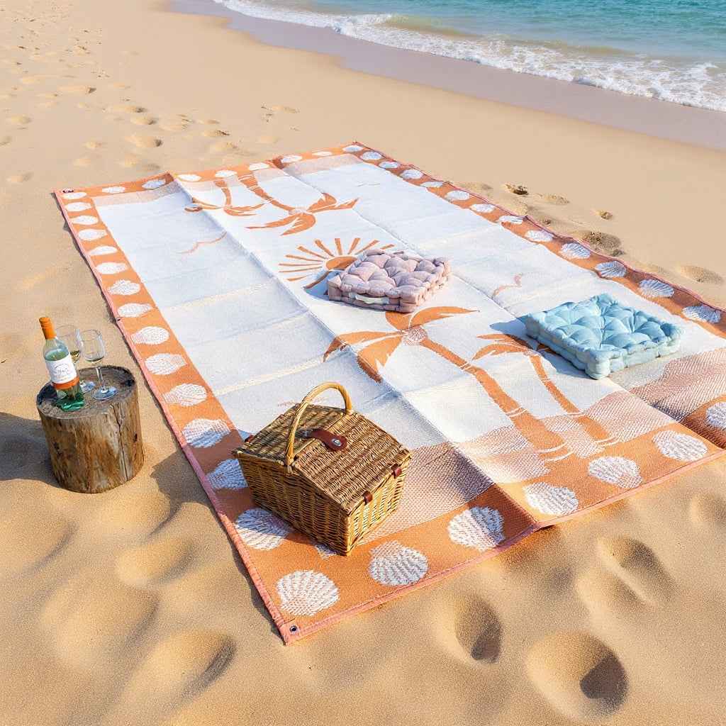Recycled outdoor beach mat with palm print, picnic basket, cushions, and wine on sandy shore