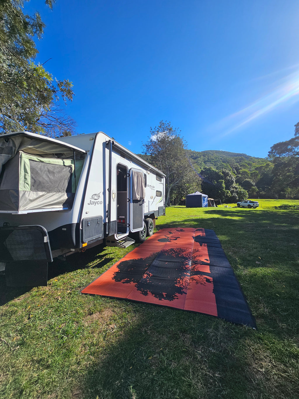 Red and black recycled outdoor mat beside caravan on grassy campsite under blue sky