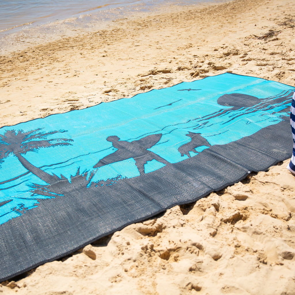 Blue and gray recycled outdoor mat with palm tree design on sandy beach, perfect for beach days and picnics.