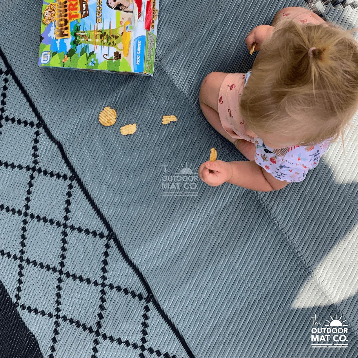 Child sitting on recycled outdoor mat with geometric pattern, eating snacks near kids' game box