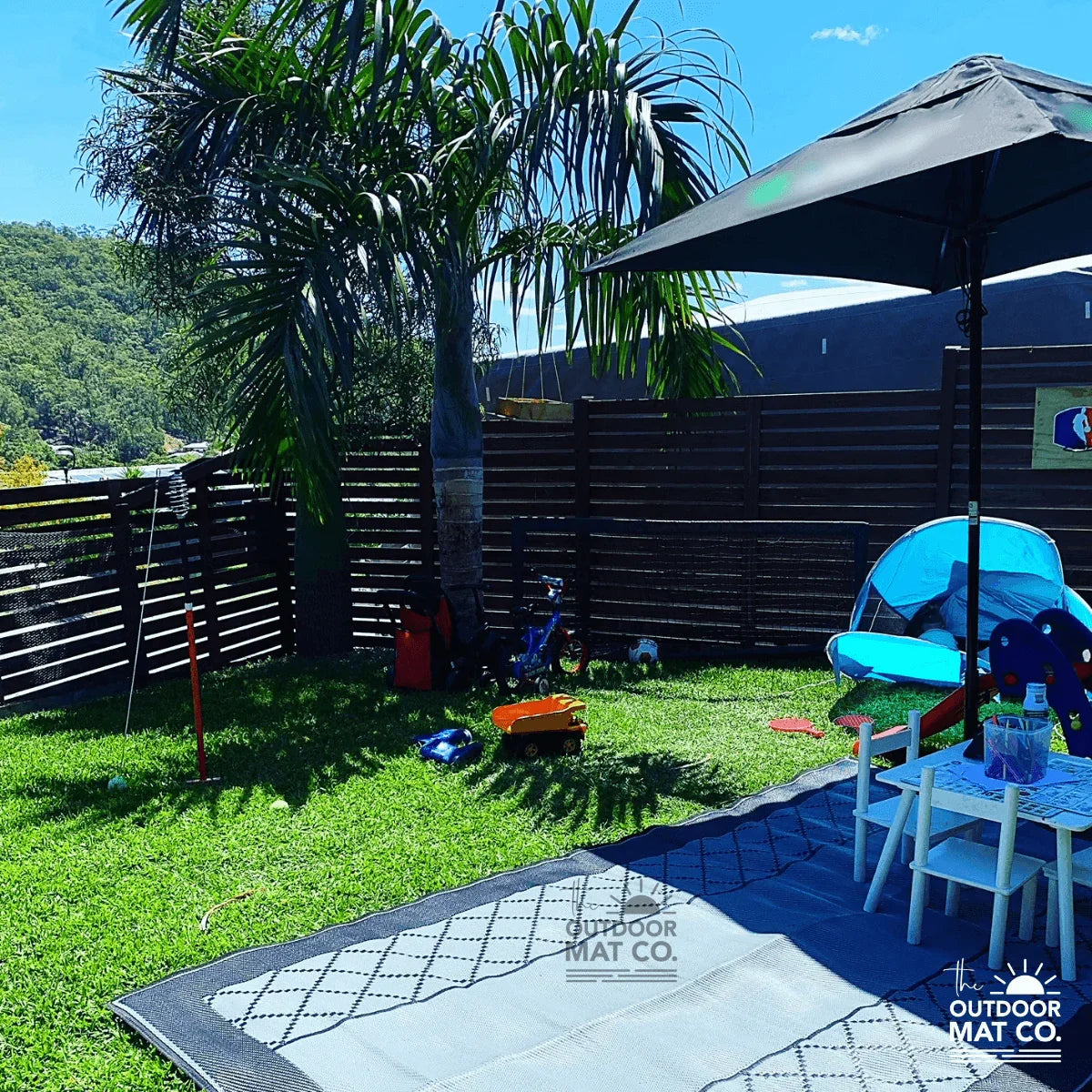 Recycled outdoor mat on grass under umbrella, perfect for picnics, camping, or beach days with kids' toys nearby