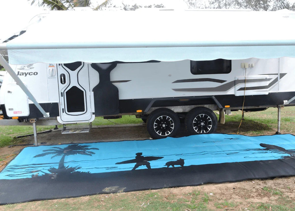 Blue beach-themed recycled outdoor mat with palm tree and surfer design, used under caravan awning for camping.