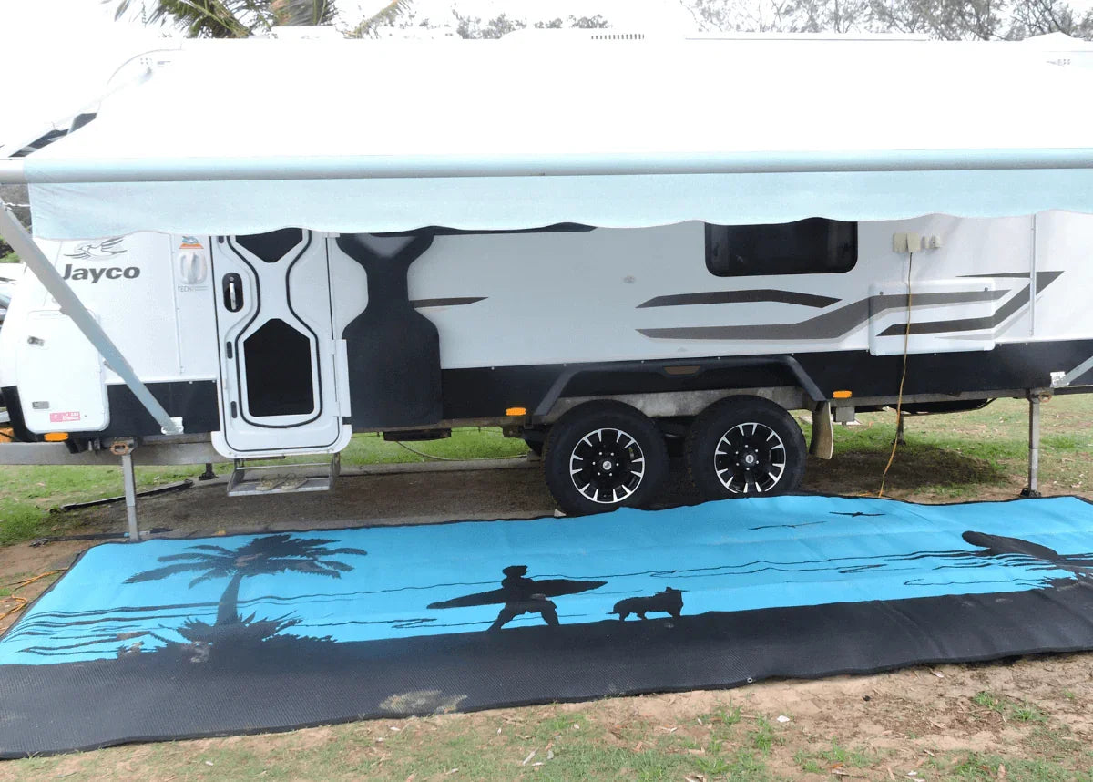 Blue beach-themed recycled outdoor mat with palm tree and surfer design, used under caravan awning for camping.