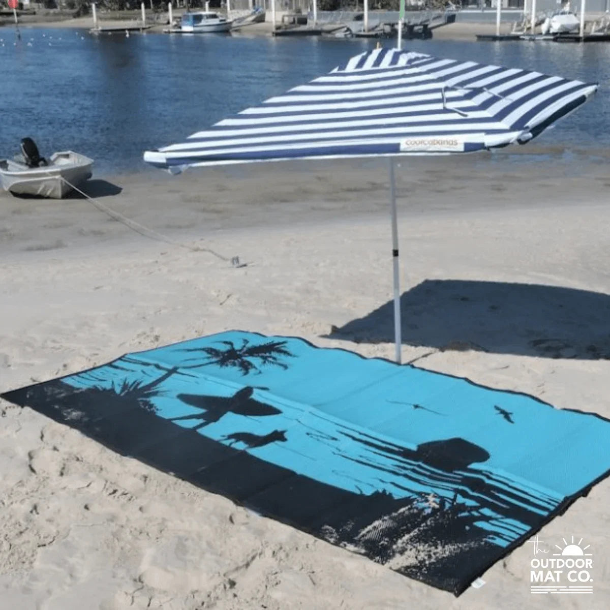 Blue and black beach mat with palm tree design under striped umbrella on sandy shore