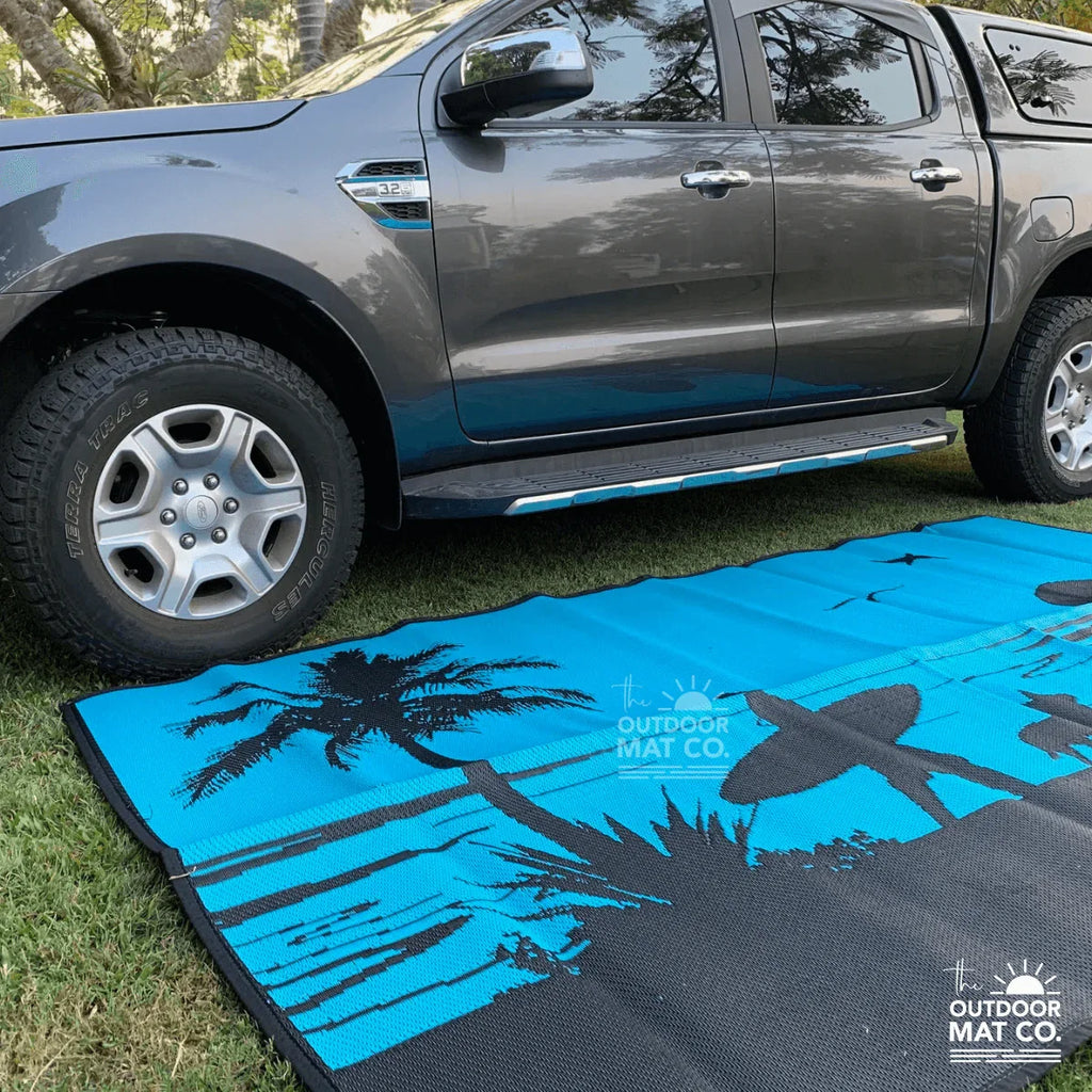 Blue beach mat with palm trees and surfboard design, laid on grass beside a pickup truck for outdoor adventures.
