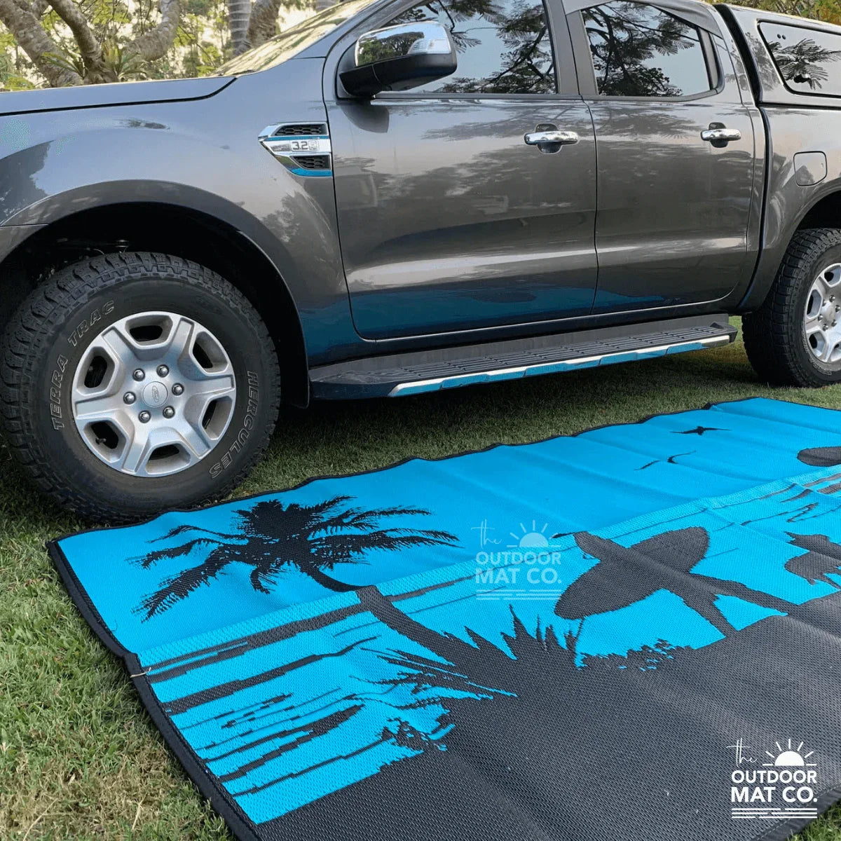 Blue beach mat with palm trees and surfboard design, laid on grass beside a pickup truck for outdoor adventures.