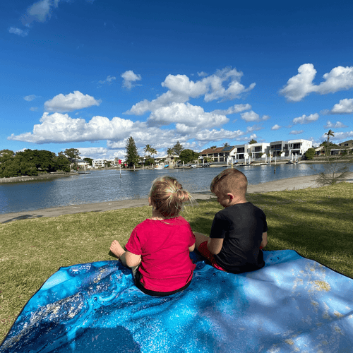 Kids sitting on blue ocean-themed recycled outdoor mat by water, perfect for picnics and beach days
