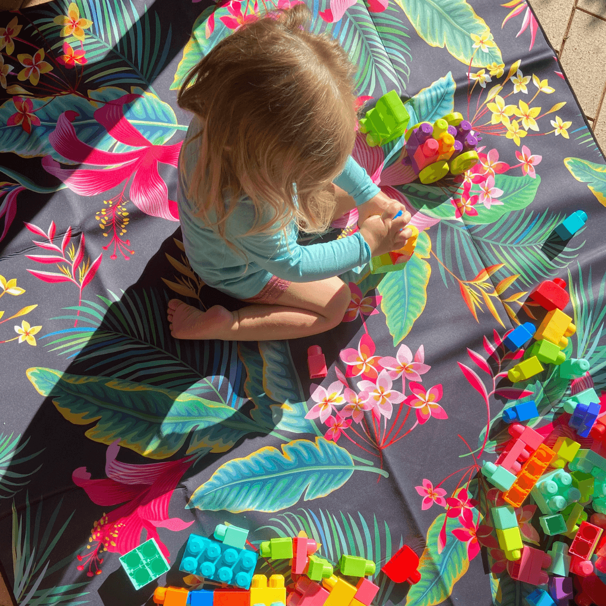 Child playing with colorful blocks on vibrant tropical print outdoor mat, perfect for picnics and beach days.