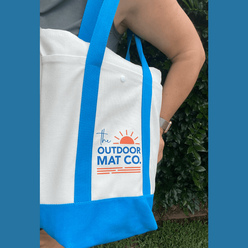 White and blue tote bag with The Outdoor Mat Co. logo, held outdoors near greenery