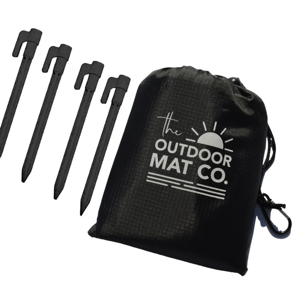 Black drawstring bag with 'The Outdoor Mat Co.' logo and four black ground stakes for securing outdoor mats